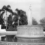 A black and white image of a fountain with a statue surrounded by palm trees and greenery.