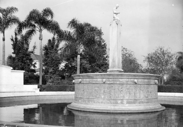 A black and white image of a fountain with a statue surrounded by palm trees and greenery.