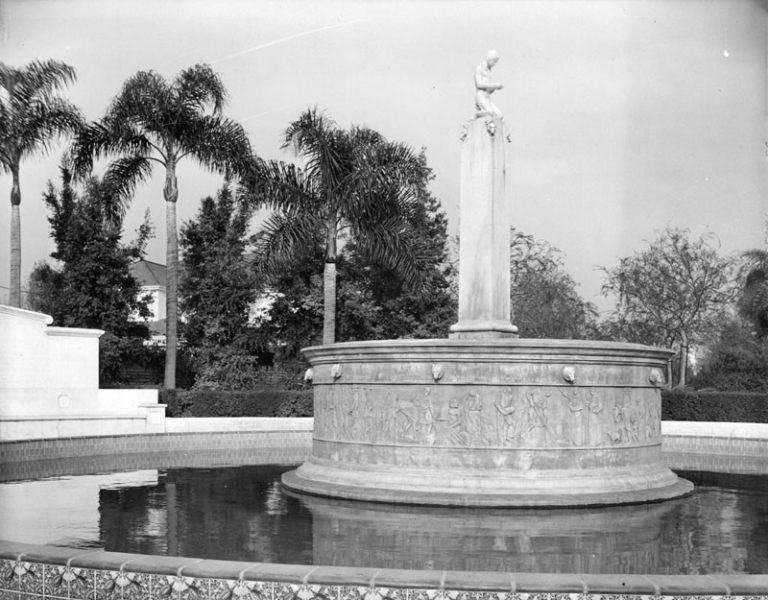 A black and white image of a fountain with a statue surrounded by palm trees and greenery.
